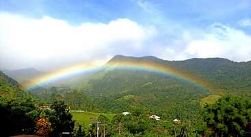 Un arco iris de vivos colores se extiende con elegancia sobre las exuberantes montañas verdes y las casas dispersas, bajo un cielo azul brillante salpicado de nubes blancas y esponjosas, creando un paisaje tranquilo y lleno de vida.