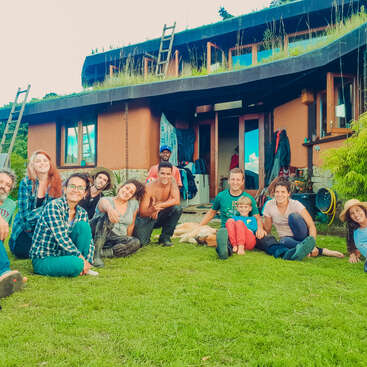 Das Bild zeigt eine Gruppe von Menschen, die im Gras vor einem Haus mit Flachdach sitzen, umgeben von einer ruhigen und natürlichen Umgebung.