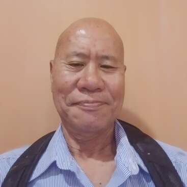 A bald man is smiling gently at the camera, wearing a blue striped shirt and black jacket. The background is plain light peach, creating a warm ambiance.