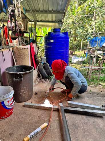 A person wearing a red headscarf is welding metal pipes outdoors near a blue water tank, surrounded by various tools and equipment in a busy workspace.