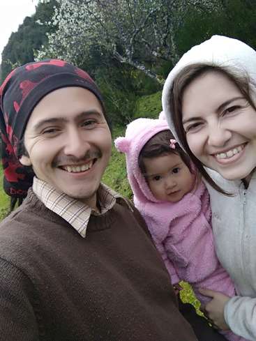 La imagen muestra a una familia de tres miembros posando para un selfie en un campo cubierto de hierba con árboles al fondo, todos ellos con ropa de abrigo.