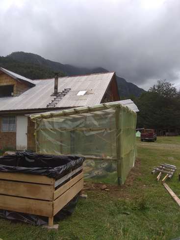 La imagen muestra una casa rústica con un invernadero y un huerto elevado, con un paisaje montañoso de fondo y un cielo nublado.