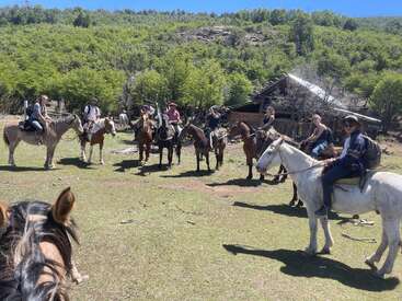 Un groupe de personnes à cheval est rassemblé dans une clairière ensoleillée et herbeuse. Elles sont entourées d'arbres verts, avec une cabane rustique en bois à proximité.