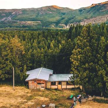 L'image représente une cabane rustique nichée dans une forêt, avec un groupe de personnes rassemblées à l'extérieur, entourées d'arbres et de montagnes en arrière-plan.
