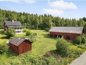 Dieses Bild zeigt eine friedliche ländliche Szene mit einem roten Bauernhaus, zwei roten Scheunen, grünem Gras, üppigen Bäumen, klarem Himmel und einer Schotterstraße in der Nähe.