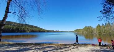 Eine friedliche Szene am Seeufer unter strahlend blauem Himmel, umgeben von immergrünen Wäldern. Drei Menschen genießen das sandige Ufer, während das ruhige Wasser die friedliche Umgebung widerspiegelt.