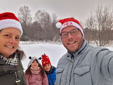 Eine vierköpfige Familie lächelt draußen in einer verschneiten Landschaft, trägt festliche Mützen und Winterkleidung und genießt gemeinsam einen fröhlichen Moment in der Weihnachtszeit.