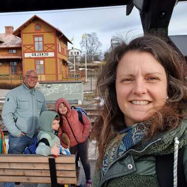 Eine vierköpfige Familie lächelt an einem Bahnhof, im Hintergrund ein gelbes Gebäude mit der Aufschrift „Ockelbo“; sie sind für das kühle Wetter warm angezogen.