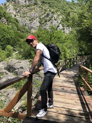 The image depicts a man standing on a wooden bridge, leaning on the railing, with a backpack on his back, set against a mountainous backdrop with trees and rocks.