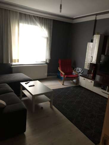 The image depicts a dimly lit living room with a couch, coffee table, TV, and red chair, featuring dark walls and a large window with white curtains.