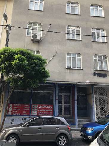 The image depicts a gray building with white-framed windows, featuring a tree and parked cars in front, alongside a storefront with red signs in an unknown language.