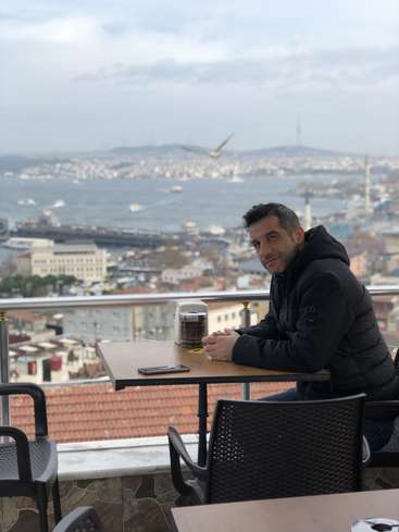 The image depicts a man sitting at a table on a balcony overlooking a cityscape, with a scenic view of the water and mountains in the background.