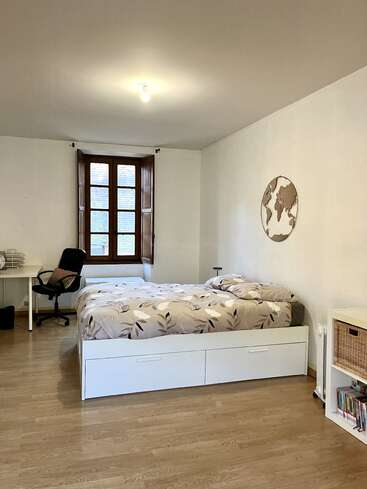 Cette chambre comprend un lit double avec une parure de lit à motifs, une grande fenêtre en bois, un bureau avec une chaise, une carte du monde accrochée au mur, des meubles blancs et un parquet en bois clair.