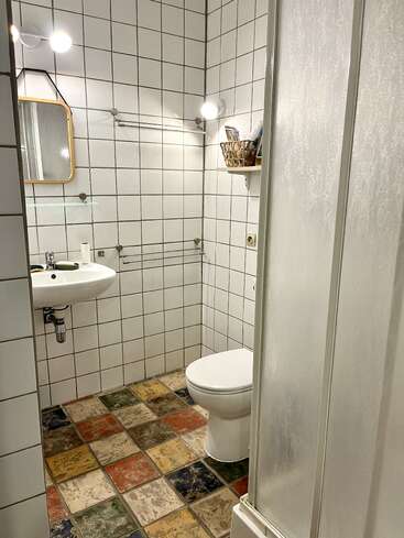 Cette salle de bains est dotée de murs carrelés blancs, d'un sol aux motifs colorés, de toilettes, d'un petit lavabo, d'un miroir, d'une étagère murale, d'un panier et d'une porte de douche en verre.
