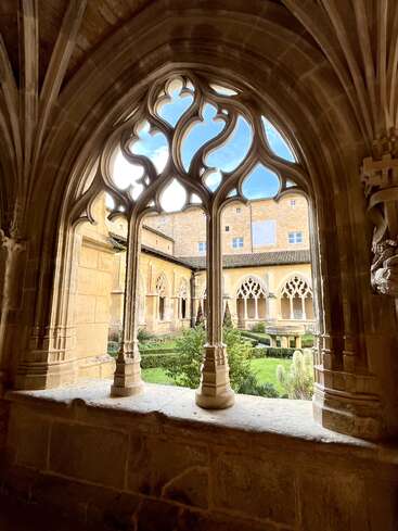 Une magnifique fenêtre en arc de style gothique encadre la cour sereine d'un monastère. Les entrelacs de pierre ouvragés, la végétation luxuriante et les murs dorés créent une atmosphère de paix et de beauté historique.