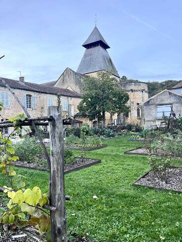 Une maison de pierre au charme d'antan, avec un haut toit pointu, se dresse derrière un jardin luxuriant où l'on trouve une pelouse verte, des treillis en bois, des parterres de fleurs et une végétation abondante.