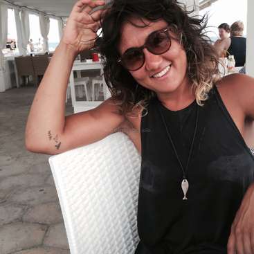 A imagem mostra uma mulher com cabelos escuros e uma camiseta preta, sentada em uma cadeira branca, com uma tatuagem no braço e um pingente em forma de peixe no pescoço.