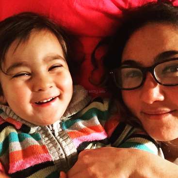 L'image montre une femme et un enfant allongés sur un lit aux draps rouges, tous deux souriant à l'appareil photo. La femme porte des lunettes et a les cheveux noirs.