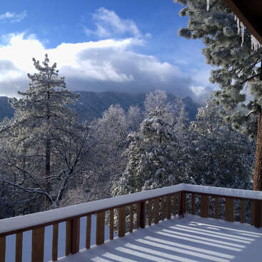 A imagem retrata uma cena serena de inverno em um deck, com árvores cobertas de neve e montanhas à distância, em um céu azul com nuvens brancas.