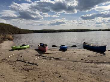 Vier farbenfrohe Kajaks und Kanus liegen auf einem sandigen Seeufer unter einem teilweise bewölkten Himmel, mit ruhigem Wasser und einem baumbestandenen Horizont in der Ferne.