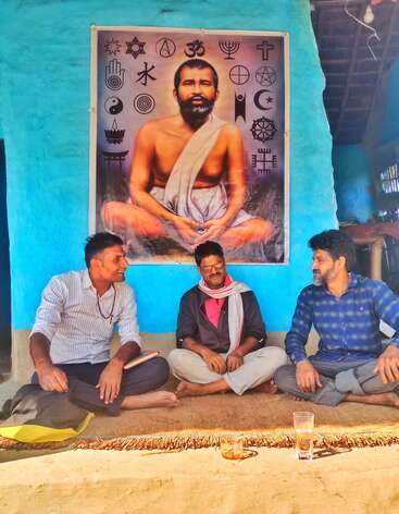Three men sit cross-legged on a mat, conversing happily. Behind them is a large poster of a meditating saint surrounded by various religious symbols.