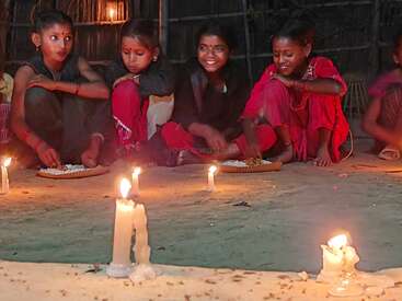 Four girls sit cross-legged on the floor, enjoying food from plates. Candles illuminate their faces, creating a warm, festive, and joyful atmosphere at night.