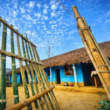 A vibrant rural scene showing a thatched-roof blue house, bamboo gates, scattered pots, under a bright blue sky dotted with fluffy clouds, radiating rustic charm.
