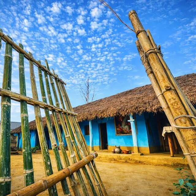 A vibrant rural scene showing a thatched-roof blue house, bamboo gates, scattered pots, under a bright blue sky dotted with fluffy clouds, radiating rustic charm.