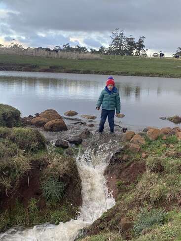 Um menino com um gorro vermelho está ao lado de um pequeno riacho que sai de um lago, cercado por campos gramados e pedras em um dia nublado.