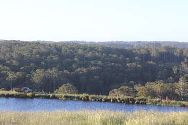 A imagem mostra uma paisagem tranquila com uma colina arborizada, um lago calmo em primeiro plano, uma caminhonete e duas pessoas ao longe.