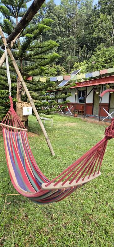 Eine bunte Hängematte ist zwischen Holzstangen auf grünem Gras aufgespannt. Bäume, Gebetsfahnen, ein rustikales Gebäude und Stühle im Freien schaffen eine friedliche Atmosphäre.