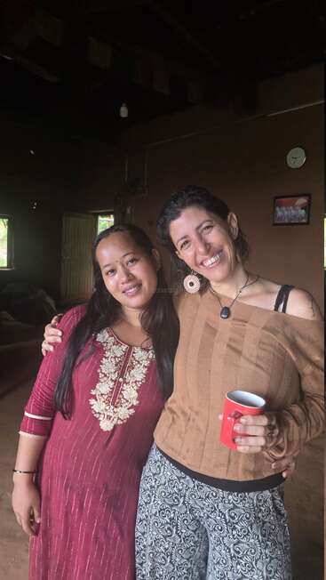 Zwei Frauen stehen lächelnd und dicht beieinander in einem Haus. Die eine trägt eine rote traditionelle Kurta, die andere einen braunen Pullover und hält einen roten Becher, der Wärme und Freundschaft ausstrahlt.