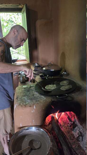 Ein Mann bereitet Fladenbrote auf einem traditionellen Holzofen in einer rustikalen Küche zu, während das natürliche Licht durch ein offenes Fenster neben ihm fällt.