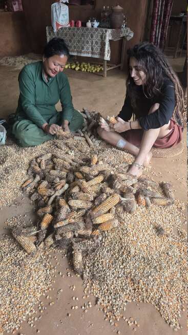 Zwei Frauen sitzen auf dem Boden und schälen fröhlich gemeinsam Maiskolben. Sie sind von einem rustikalen Raum umgeben, in dem Obst und Haushaltsgegenstände auf einem Tisch im Hintergrund stehen.