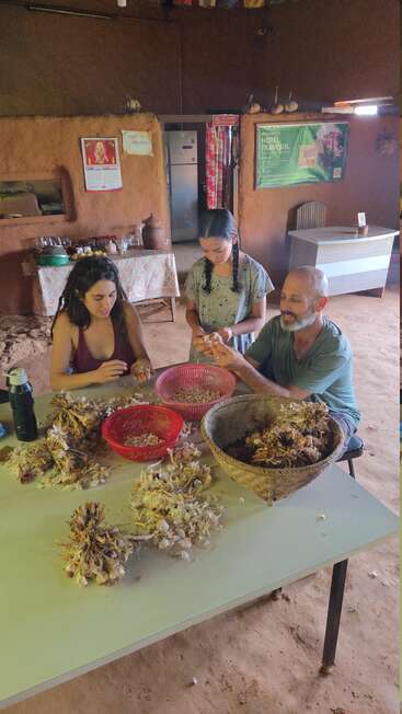 Drei Personen sitzen um einen Tisch in einem rustikalen Raum und schälen oder trennen Knoblauchzwiebeln, während Körbe und Küchenutensilien auf dem Tisch und im Hintergrund verstreut sind.