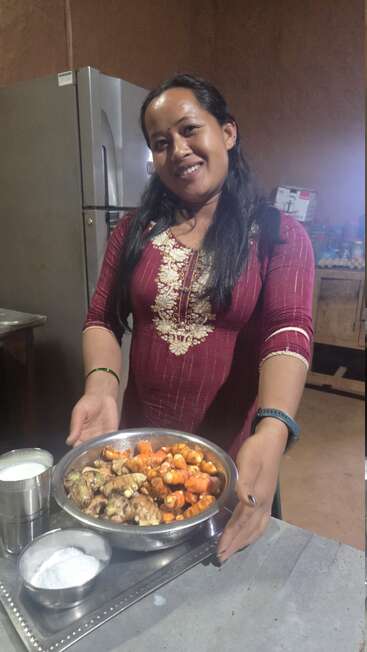 Eine lächelnde Frau in einem kastanienbraunen ethnischen Kleid hält ein Tablett mit frischer Kurkuma, Ingwer, Salz und einem Glas Milch in einer rustikalen Küche.