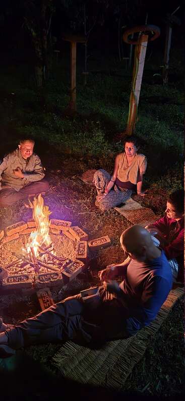 Vier Menschen sitzen nachts auf Matten um ein kleines Lagerfeuer und genießen die Wärme und das Licht. Bäume und Holzskulpturen stehen ruhig im Hintergrund.
