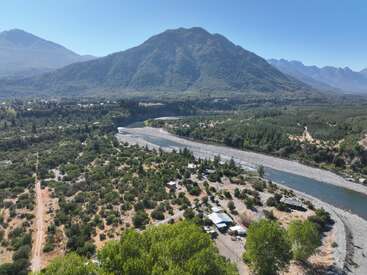 A river winds through a lush, green valley surrounded by dense forests and mountains. Small houses and pathways dot the landscape under a clear blue sky.