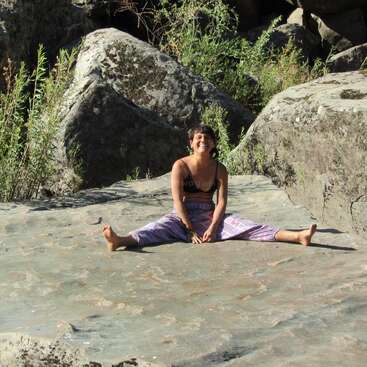 Una mujer sentada en una gran roca, vestida con un top negro y rosa con pantalones a juego, en una pose de piernas abiertas, con las manos entrelazadas entre las piernas.