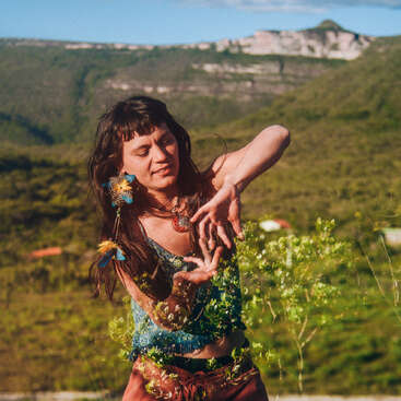 A woman dances gracefully outdoors, blending with nature. Her arms are elegantly posed, adorned with feathers, and she appears joyful amidst the lush green landscape and mountains.