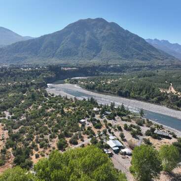A river winds through a lush, green valley surrounded by dense forests and mountains. Small houses and pathways dot the landscape under a clear blue sky.