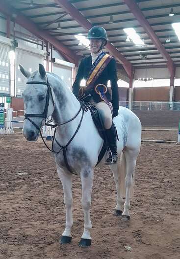 Un jinete sonriente, con una banda de campeón y un casco, se sienta orgulloso sobre un caballo blanco dentro de una pista cubierta, listo para una competición de salto ecuestre.