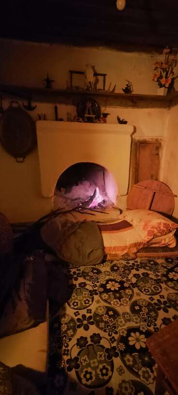 Une pièce rustique et chaleureuse, avec une cheminée allumée, un tapis à motifs, des coussins et des couvertures. Des étagères accueillent des objets décoratifs, créant une atmosphère chaleureuse, accueillante et nostalgique.