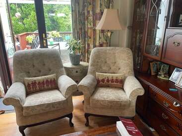 Two beige armchairs with decorative cushions face the room. A lamp, side table, and plant sit between them. Wooden furniture and large windows complete the cozy scene.