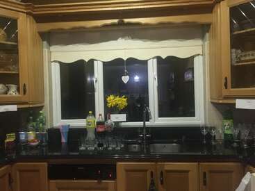 This cozy kitchen has wooden cabinets, a black countertop, glassware, soft drinks, colorful straws, a window with blinds, and yellow flowers in a vase by the sink.