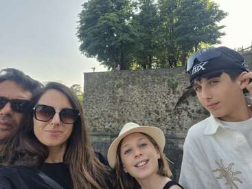 Une famille de quatre personnes prend un selfie en plein air. Tous sont vêtus de tenues d'été et portent des lunettes de soleil ; ils sourient devant un vieux mur de pierre et des arbres à la verdure luxuriante.