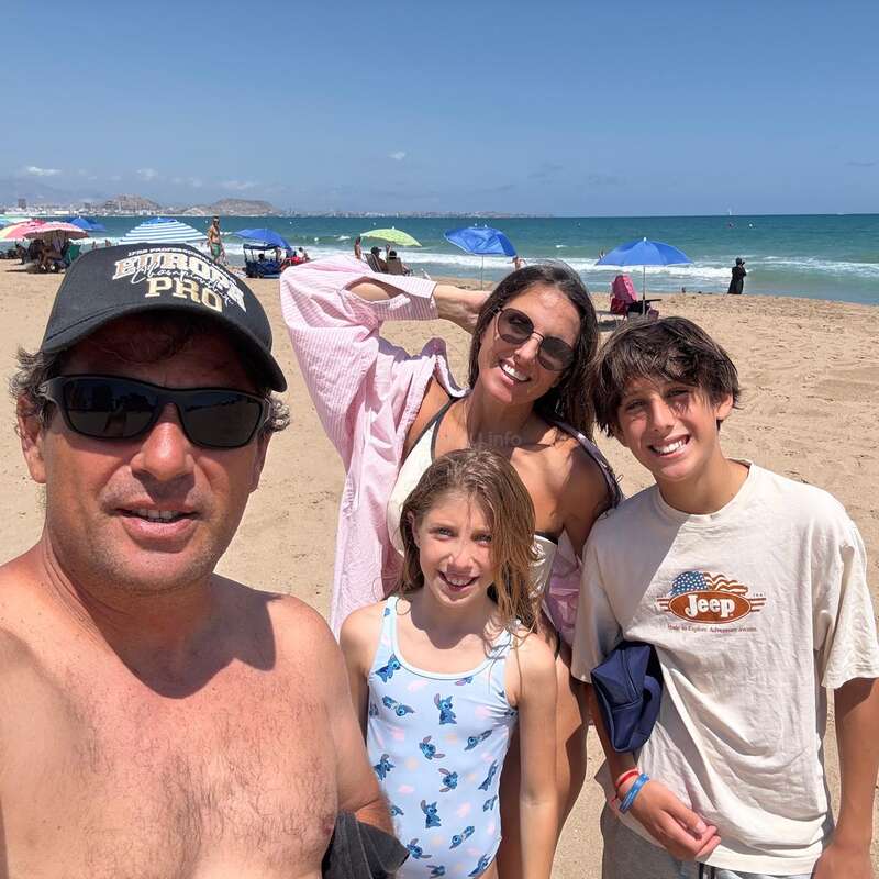 Une famille de quatre personnes pose en souriant sur une plage ensoleillée. Ils portent des vêtements d'été et des lunettes de soleil. On aperçoit des parasols colorés et l'océan en arrière-plan.