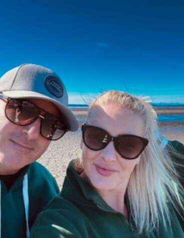 Un hombre y una mujer con gafas de sol se hacen un selfie en un día soleado en la playa, vestidos con ropa informal, disfrutando juntos del hermoso cielo azul.