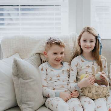 Two children sit on a light-colored couch, wearing matching bunny pajamas. The girl holds an Easter basket. Both smile warmly in a softly lit, cozy room.