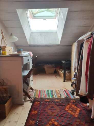 Um quarto aconchegante no sótão com paredes de madeira, uma claraboia, tapetes coloridos, uma escrivaninha com cadeira, luminária, cabideiro e cestos de tecido para armazenamento.